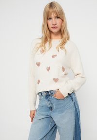 Helles beiges Sweatshirt mit rosa Herzmustern, Rundhalsausschnitt, gerippte Bündchen, kombiniert mit hoch taillierten blauen Jeans. Weicher Stoff.
