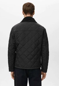 Giacca nera trapuntata con colletto tradizionale, caratterizzata da motivi a diamante e dettagli cuciti, abbinata a pantaloni di denim scuro.