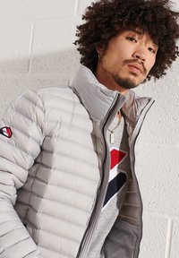 Veste doudoune gris clair avec texture matelassée, col montant et fermeture à zip. Portée par-dessus une chemise grise ornée de motifs rouges et bleus marine.