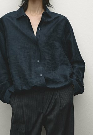 Personne portant une chemise noire ample texturée à boutons avec les manches retroussées et un pantalon noir à fines rayures, les mains dans les poches.
