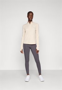 Lys beige hettegenser med glidelås og lange ermer, samt mørkegrå leggings, begge laget av glatt, elastisk stoff; hvit logo-detalj; kombinert med hvite joggesko.
