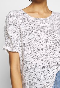 Blusa blanca con pequeños puntos negros dispersos, mangas cortas abullonadas, combinada con jeans de denim azul claro, mostrando el cuello y la parte superior del brazo.