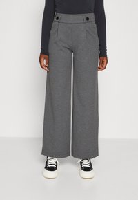 Osoba stojí v šedých širokých kalhotách, černém dlouhém rukávu a teniskách, což vytváří ležérní, ale stylový outfit.