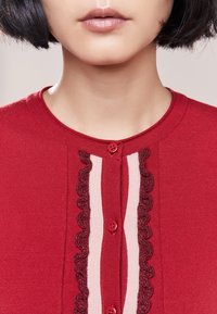 Punainen neuletakki pyöreällä kauluksella, jossa on vaaleanpunainen raita ja burguundin väriset aaltoilevat yksityiskohdat edessä, kiinnitetty punaisilla nappeilla.