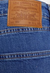 Hnědá kožená záplata na modrých riflích, označená jako „Levi Strauss & Co“ s nápisy „Lot 502,“ „W 31 L 32“ a cvočky jako akcenty.