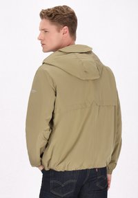 Jachetă bej tip windbreaker cu glugă, dotată cu mansete elastice și tiv ușor strâns, surprinsă din spate pe un fundal deschis.