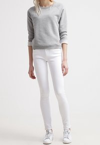 Sudadera gris de cuello redondo con borde blanco, combinada con unos vaqueros blancos ajustados y zapatillas blancas. Ropa casual y ajustada en textos suaves.