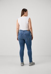 Προχειρισμένο λευκό αμάνικο μπλουζάκι και μπλε στενά τζιν, με πίσω τσέπες και δερμάτινη ετικέτα στη ζώνη, φορεμένο με γκρι αθλητικά παπούτσια.