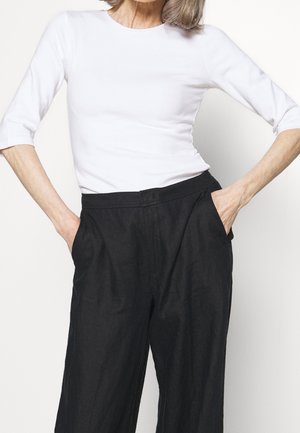 Vrouw draagt een wit shirt met driekwart mouwen en losse zwarte broek, met handen in de zakken tegen een effen lichte achtergrond.