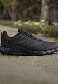 Scarpa da trail outdoor Adidas nera con suola strutturata ed etichetta Gore-Tex, posizionata su un sentiero sterrato con sfondo di foresta sfocata.