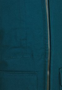 Teal-färgad tygjacka med dragkedja framtill, två stora fickor och synlig sömnad. Texturen verkar hållbar med en canvasliknande väv.