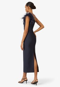 Abito aderente blu navy con dettagli in piume su una spalla, spacco laterale e design elegante. Tacchi a strappo neri completano l'outfit.