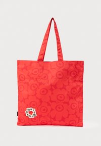 Κόκκινη τσάντα tote με δύο λαβές, διακοσμημένη με έντονο λουλουδένιο σχέδιο και ένα μικρό λευκό σημείο με κείμενο κοντά στην κάτω αριστερή γωνία.