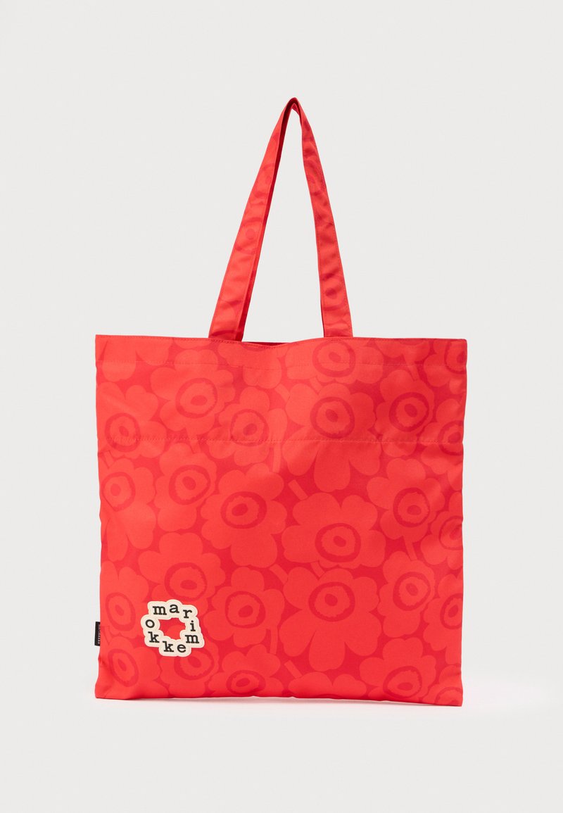 Κόκκινη τσάντα tote με δύο λαβές, διακοσμημένη με έντονο λουλουδένιο σχέδιο και ένα μικρό λευκό σημείο με κείμενο κοντά στην κάτω αριστερή γωνία.