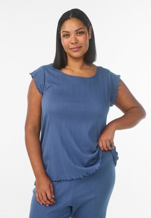 Kobieta o ciemnych, prostych włosach ubrana w niebieski prążkowany top z krótkim rękawem i pasujące spodnie, pozująca z jedną ręką na biodrze.