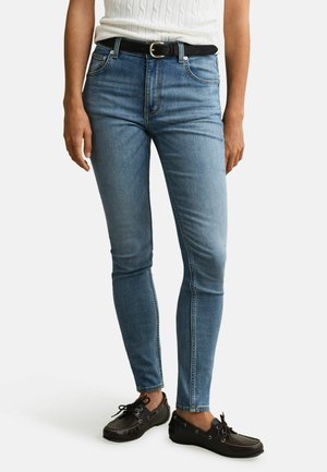 Frau trägt hellblaue Skinny-Jeans mit schwarzem Gürtel, ein weißes ärmelloses Oberteil und schwarze Leder-Mokassins, steht vor weißem Hintergrund.