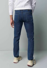 Jean en denim bleu foncé avec une coupe slim, cinq poches et une patch en cuir noir à la ceinture arrière. Porté avec des baskets de couleur claire.
