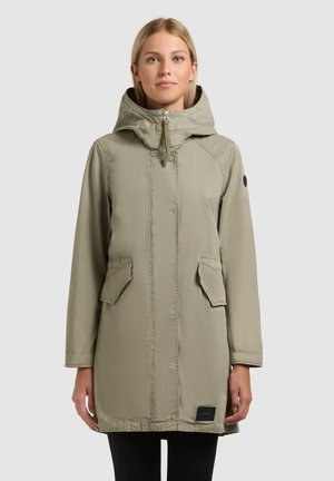 Femeie purtând o parka cu glugă de culoare verde măsliniu deschis, cu fermoar frontal și buzunare cu clape, stând pe fundal gri simplu.