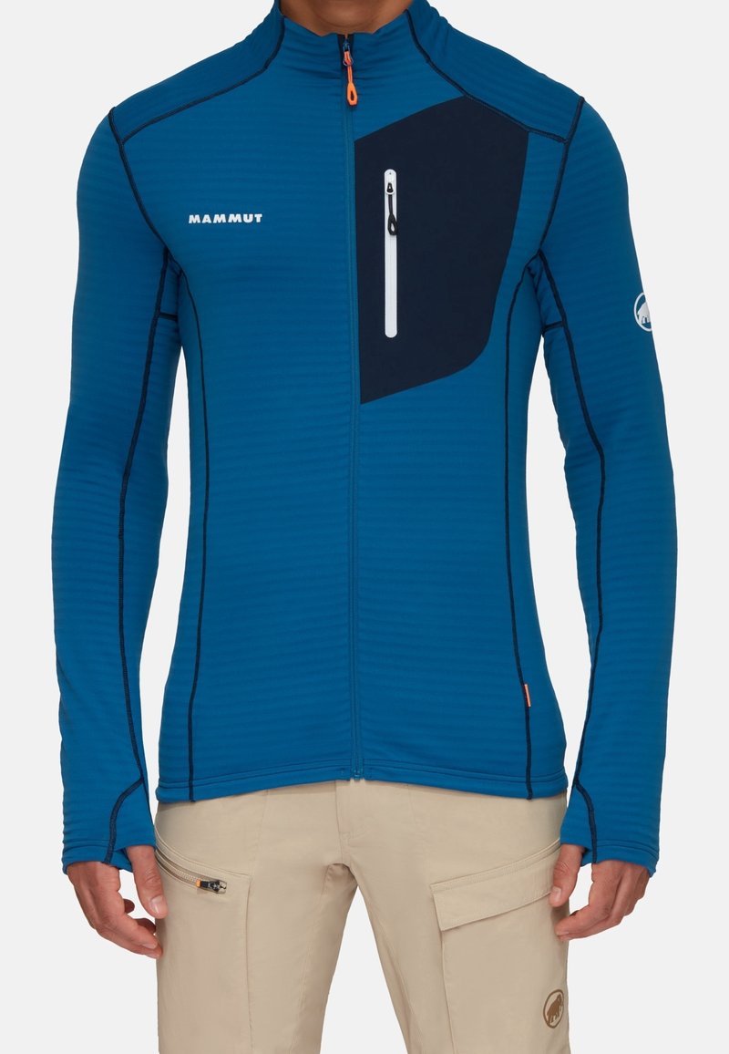 Veste zippée bleue avec texture côtelée verticale, panneau contrastant marine, poche zippée à l'avant et accents de couture noirs. Conception légère et ajustée.