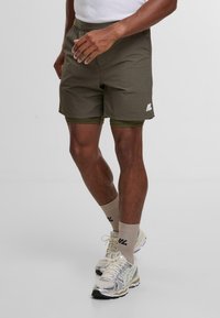 Shorts deportivos de color verde oliva con una cintura elástica, detalle de logotipo blanco, y capas de pantalones cortos interiores verde oliva, combinados con calcetines beige y zapatillas.