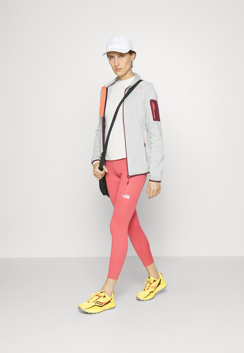 Graue Zipjacke mit orangefarbenen und roten Akzenten, weißem T-Shirt, pinken Leggings und gelben Laufschuhen. Schwarze Messenger-Tasche und weiße Mütze.