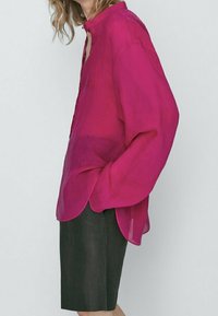 Fuchsia sheer button-up shirt with a high collar, relaxed fit, and rounded hem, paired with dark tailored shorts.