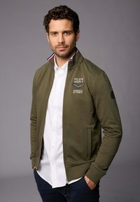 Veste zippée olive avec une texture côtelée et un col montant. Dotée d'un accent rayé blanc, rouge et bleu sur le col et de détails brodés.