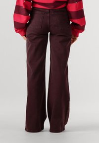 Persoon draagt een donker bordeauxrode wijde broek en een rood gestreepte lange mouwen top, staande met zijn/haar rug naar de camera tegen een effen achtergrond.