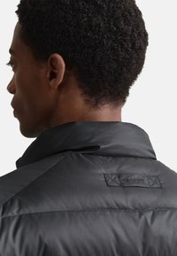 Juoda paminkštinta striukė su aukštu apykakliu, gale siuvinėtas lipdukas. Lygi tekstūra su geometriniais siūlių raštais.