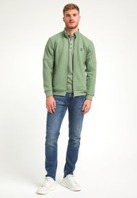 Chaqueta verde texturizada con cuello alto, llevada sobre un polo verde claro, combinada con jeans azules y zapatillas blancas.