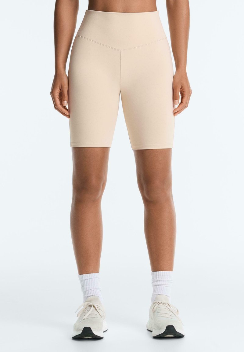 Personne portant un short de vélo beige taille haute, des chaussettes blanches et des chaussures de sport blanches, debout devant un fond clair uni.