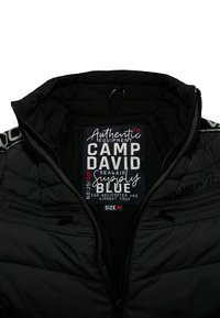 Schwarze Pufferjacke mit einem Reißverschlusskragen, die ein Etikett innen enthält, auf dem Marken- und Größeninformationen angegeben sind. Strukturierte, isolierte Ausführung.