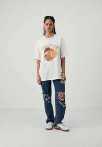 Vit överstor t-shirt med orange grafik och texten "ORANGE," tillsammans med blå rivna jeans och vita sneakers med gröna accenter.