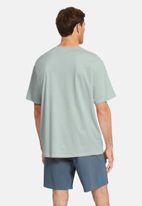 Camiseta de manga corta verde claro en tejido suave, con un corte relajado y dobladillo recto. Llevada con pantalones cortos azul marino que muestran un logotipo.