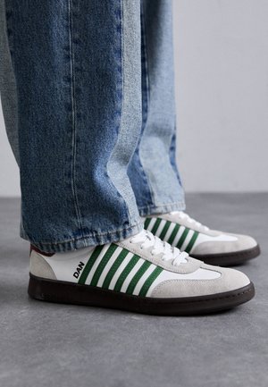 Sneaker bianco e verde realizzato in suede e pelle, con strisce verdi, lacci bianchi e suola in gomma. Abbinato a jeans blu.