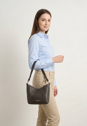 Vrouw met lichtblauw shirt en beige broek, met een zwarte Venezia-handtas over haar schouder tegen een effen achtergrond.