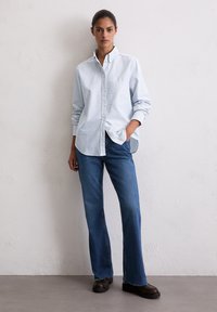 Blauw-wit gestreept overhemd met knopen en borstzak, gecombineerd met blauwe flared jeans en zwarte schoenen, voor een neutrale achtergrond.