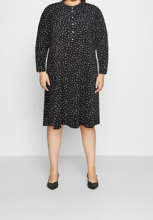 Nainen pukeutunut mustaan polvipituiseen mekkoon, jossa on valkoisia pilkkuja, pitkät hihat, edessä napit ja mustat kärjestä terävät korot, seisomassa harmaalla lattialla.