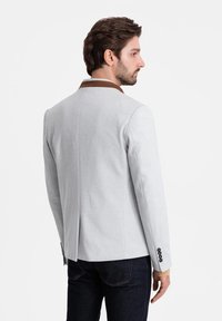 Giacca grigia chiara con colletto marrone, vestibilità sartoriale, spacco singolo sul retro e polsini con bottoni; il tessuto appare liscio e strutturato.