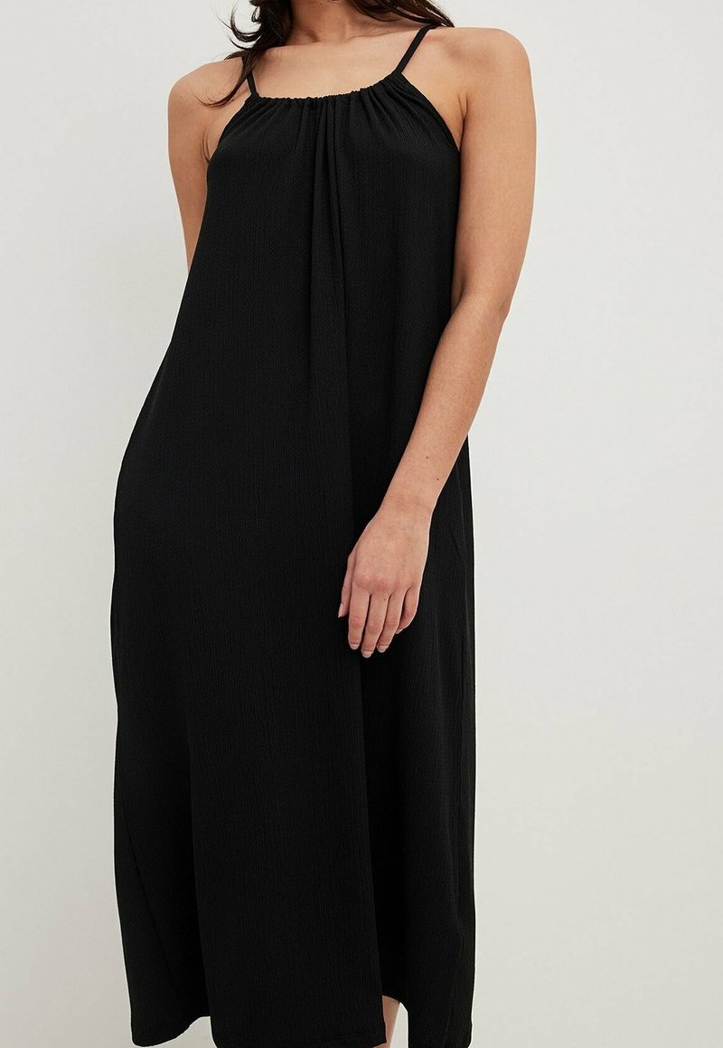 Femme portant une robe midi noire sans manches avec de fines bretelles et un décolleté froncé sur un fond clair uni.