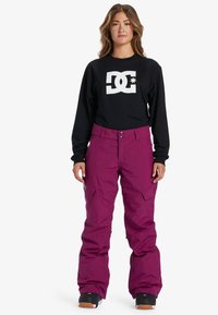 Zwarte longsleeve shirt met witte logo, gecombineerd met paarse sneeuwbroeken. De broeken hebben een losse pasvorm en meerdere zakken. Zwarte sneeuwlaarzen zijn zichtbaar.