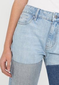 Ljusblå denimjeans med patchworkdesign; har blekta områden och mörkblå sektioner; inkluderar framfickor och metallnitar.