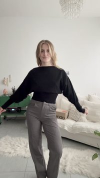 Femme aux cheveux blonds portant un pull noir et un pantalon gris debout dans un salon avec un canapé blanc et un meuble vert.
