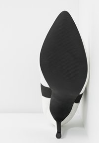 Zapato blanco de tacón alto con puntera puntiaguda, que presenta una suela texturizada negra y acentos de bandas elásticas negras alrededor de la parte media del pie.
