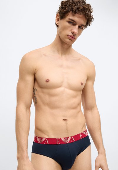 Cueca masculina azul-marinho com cintura elástica vermelha com logo. O tecido é liso, com ajuste justo, apresentando um bolso frontal definido. Sem padrões adicionais.