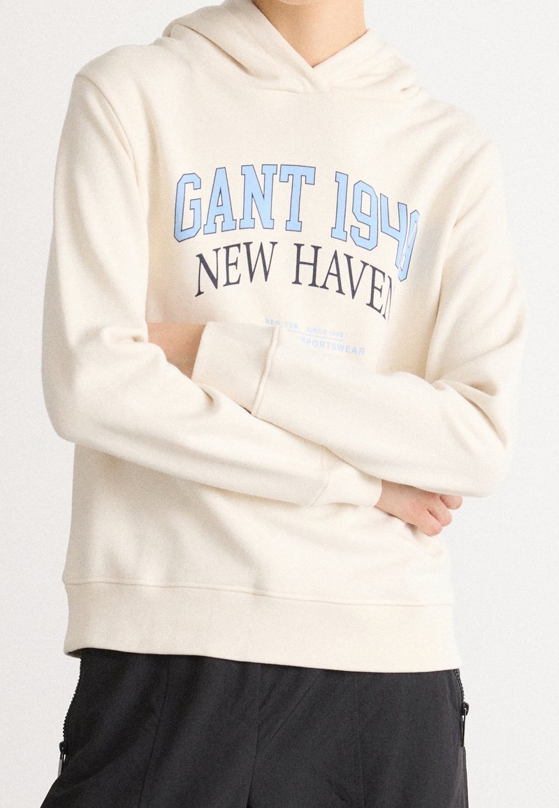 Personne portant un sweat à capuche crème avec le texte « GANT 1949 NEW HAVEN » en bleu et noir, associée à un pantalon noir, bras croisés.