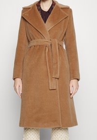 Bruine wolmixjas met een uitgesneden kraag, ceintuur in de taille en zijzakken, met een contrasterende geruite voering die zichtbaar is bij de kraag.