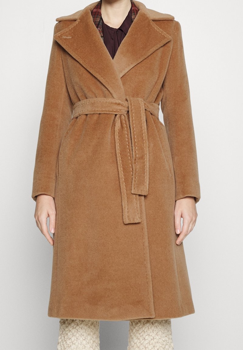 Bruine wolmixjas met een uitgesneden kraag, ceintuur in de taille en zijzakken, met een contrasterende geruite voering die zichtbaar is bij de kraag.