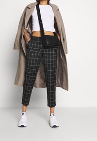 Pantalon à carreaux noir et blanc avec un top blanc court, un manteau beige et des baskets blanches. Sac bandoulière noir avec une lanière.