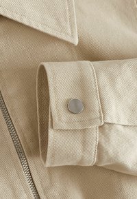 Vêtement en coton beige avec un col pointu, un bouton-pression argenté au poignet et une fermeture éclair visible avec un motif de tissu texturé.